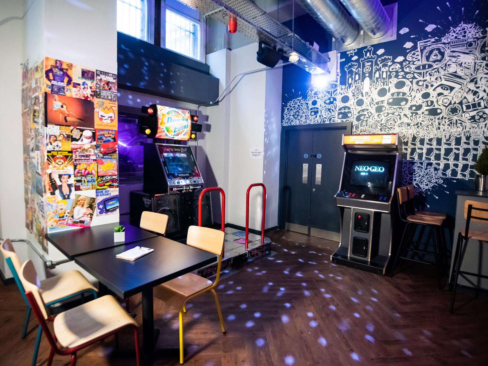 A bar with vintage arcade machines, a mural wall, and a small table with chairs at Four Quarters Newcastle.