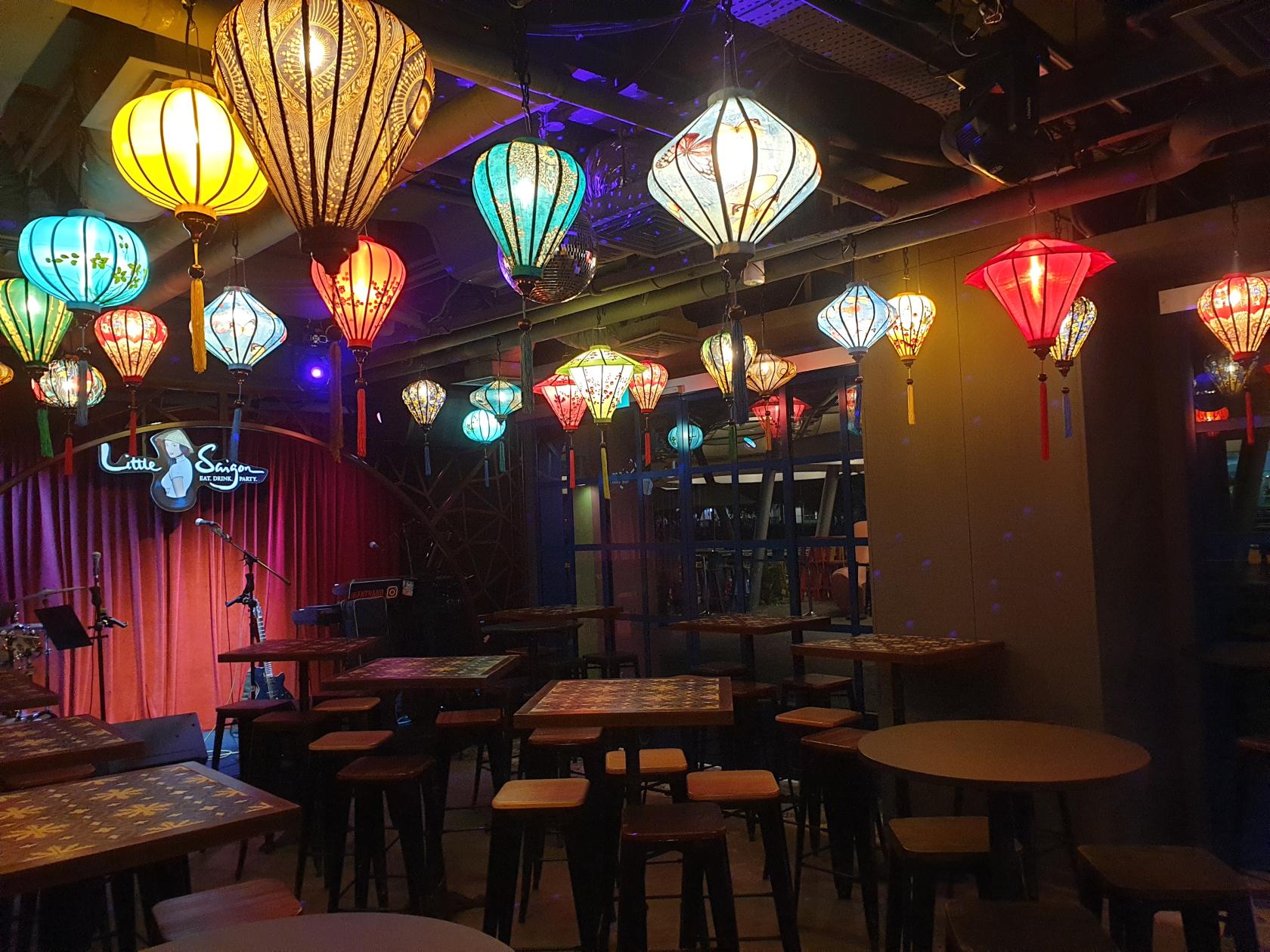 A restaurant with colorful hanging lanterns, wooden tables, and a stage with red curtains at Little Saigon.
