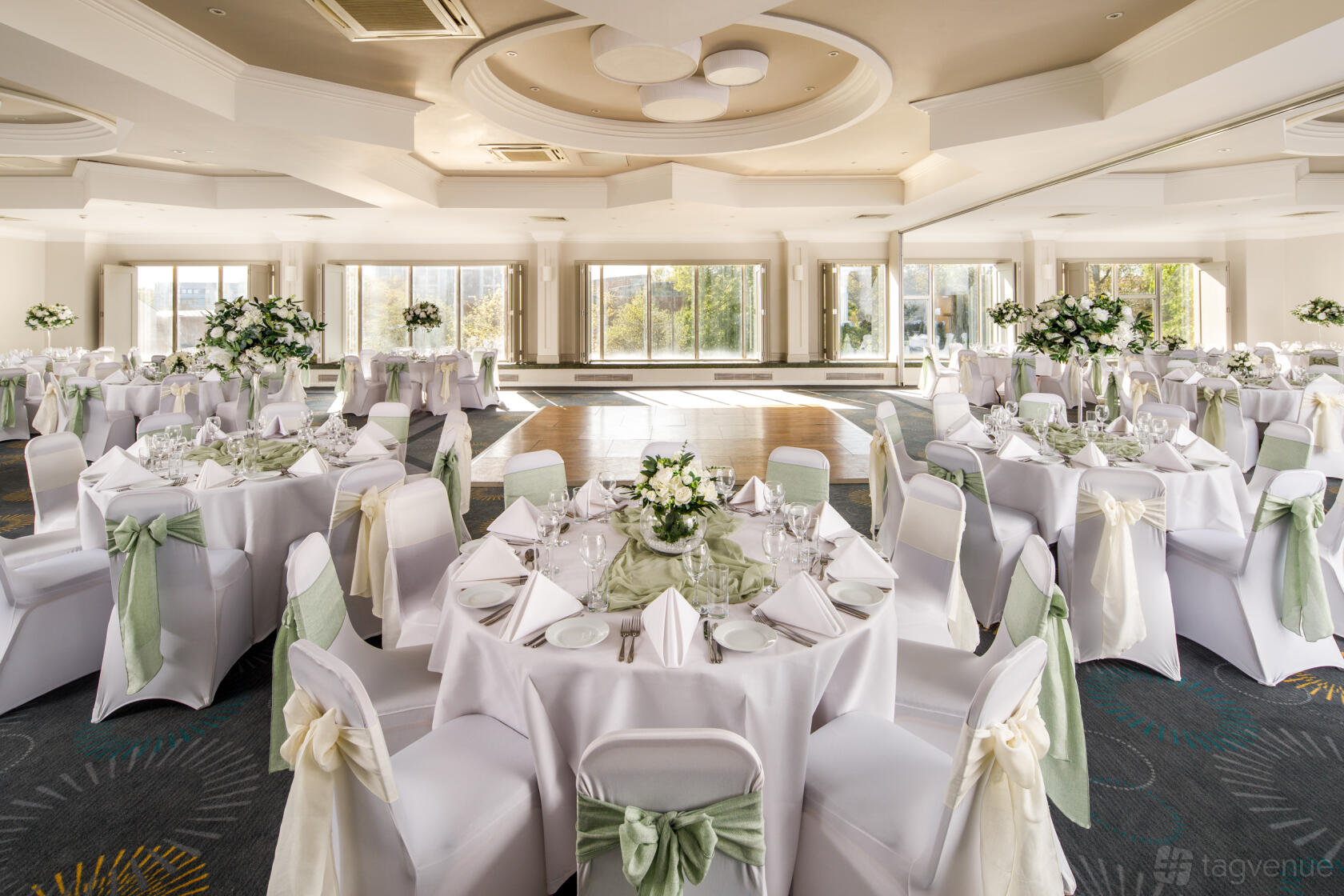 An event space with round tables, white linens, elegant chair covers, and large windows at Mercure Bedford Centre.