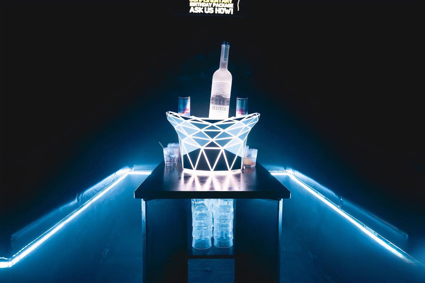 A nightclub dining room with a glowing geometric bottle holder, vodka bottle, and illuminated booth seating at Mi Casa.