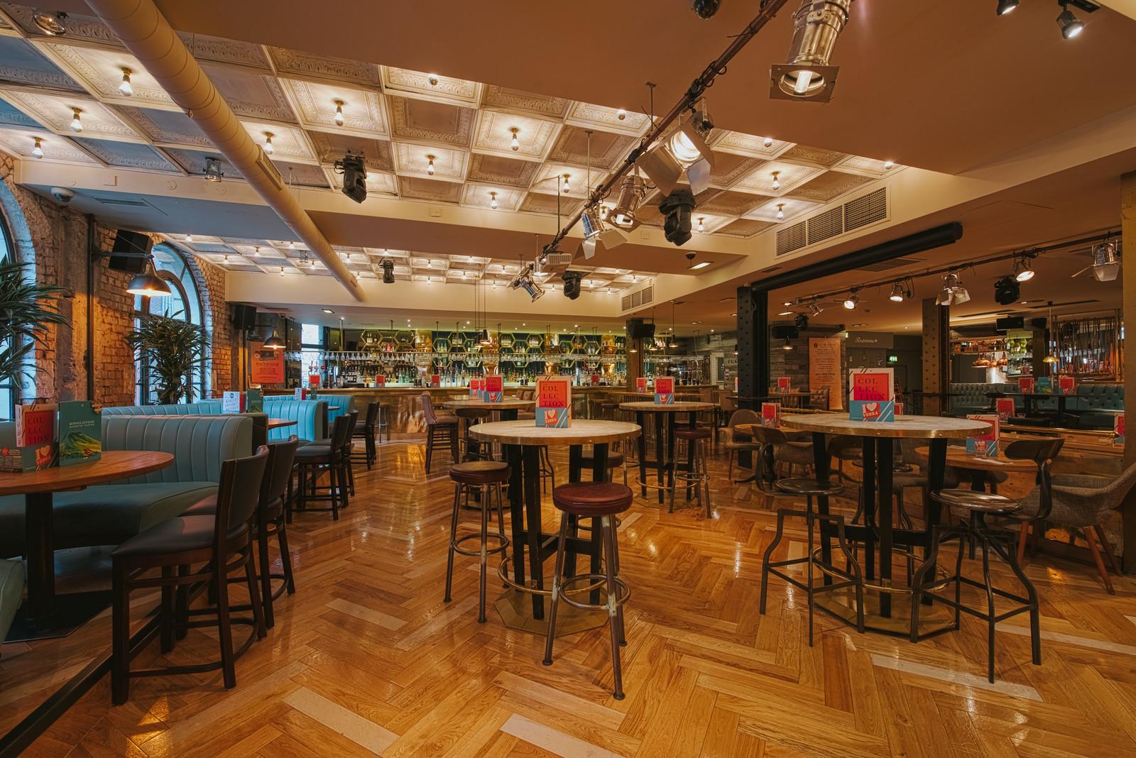 A bar with parquet wood floors, booth seating, round tables, and a mirrored back bar at Revolution Parsonage Gardens.