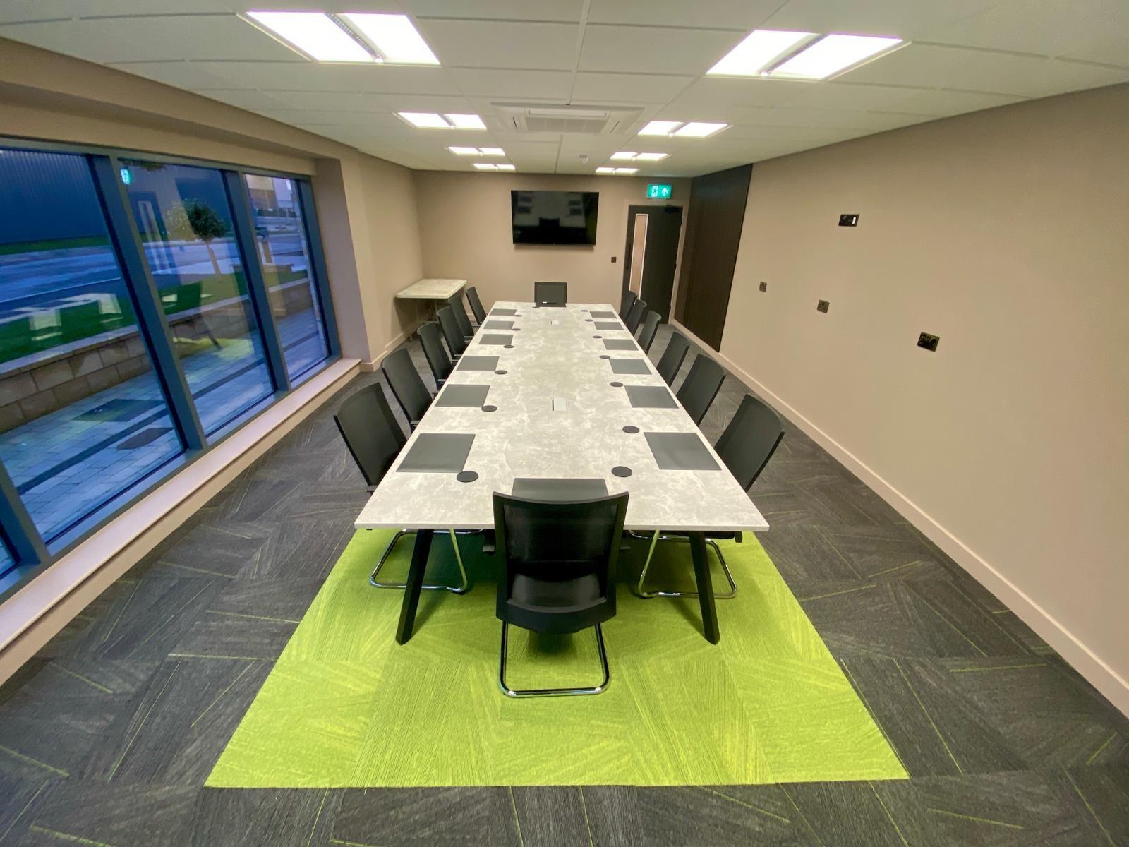 A meeting room with a long rectangular table, black chairs, large windows, and wall-mounted screen at Enterprise House.