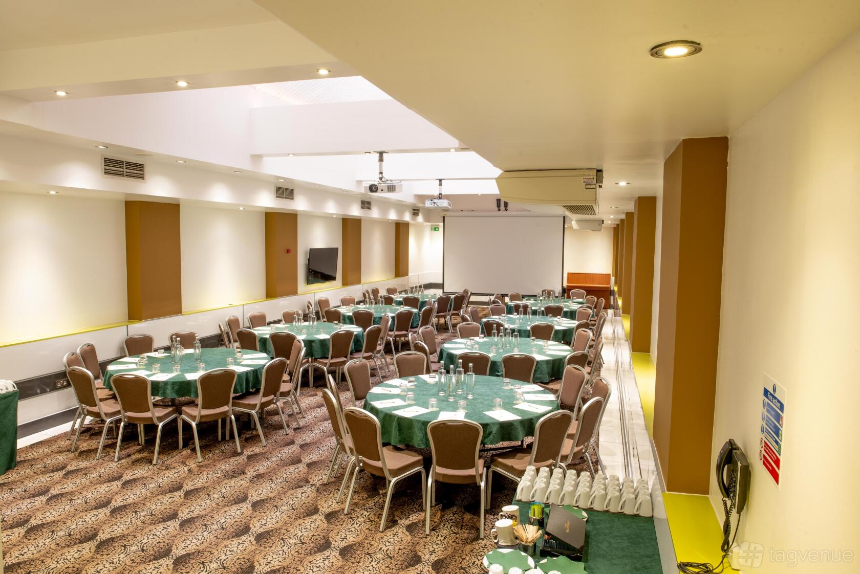A hotel hall with round tables covered in green cloths, banquet chairs, and a large projector screen at The Wesley Euston Hotel.