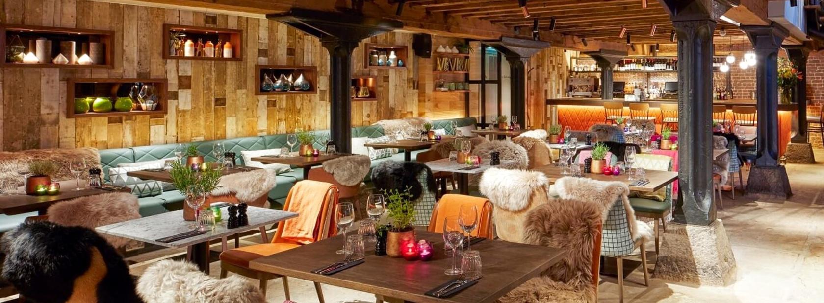 A basement bar with wooden walls, exposed beams, fur-covered chairs, and candlelit tables at The Sipping Room.