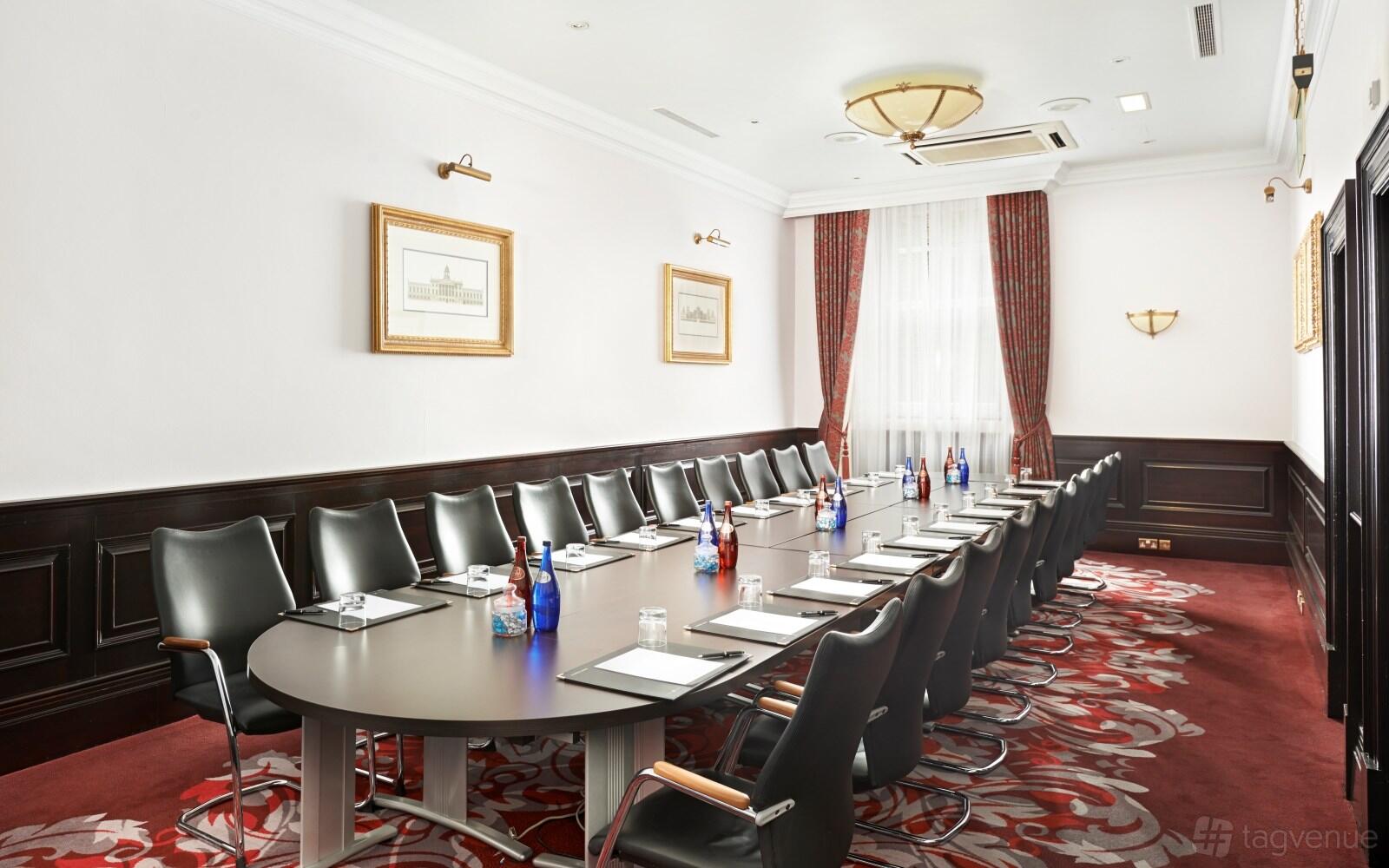 A hotel boardroom with a long oval table, black leather chairs, white walls, and red patterned carpet at Clermont Charing Cross.