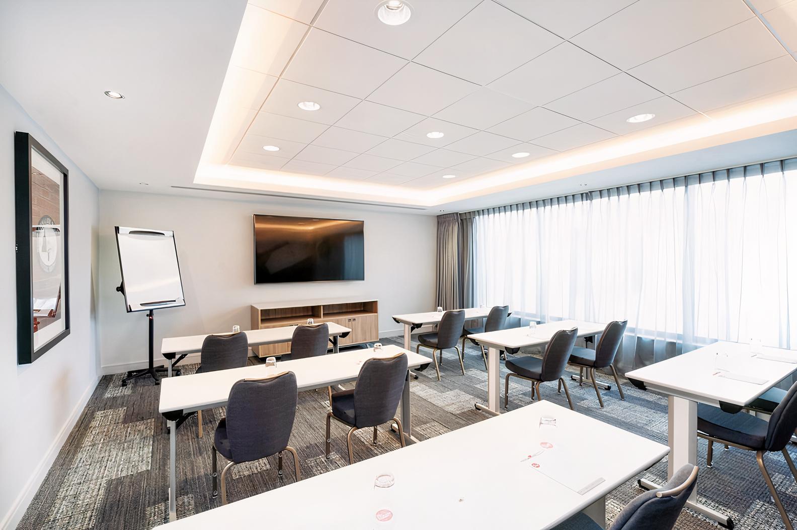 A conference meeting room with rows of tables, navy chairs, a large wall-mounted screen, and floor-to-ceiling windows at The Eastside Rooms.