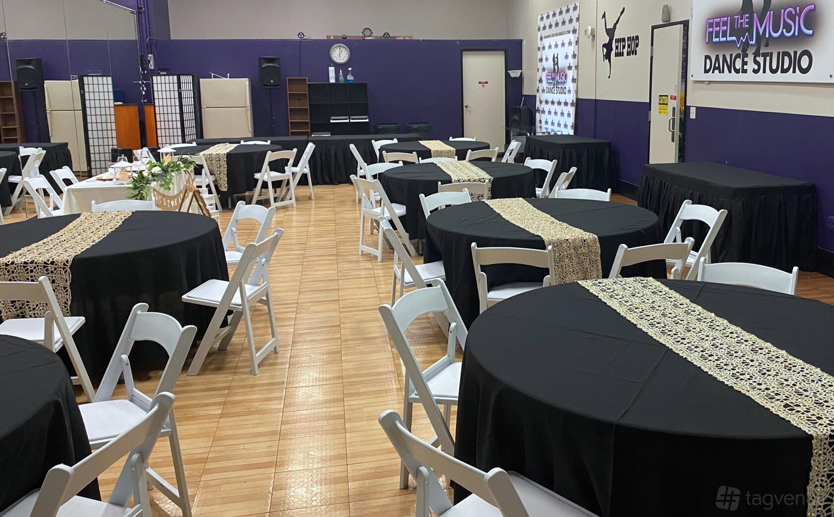 A dance studio with round tables covered in black cloths and white chairs on a wooden floor at Feel The Music Dance Studio.