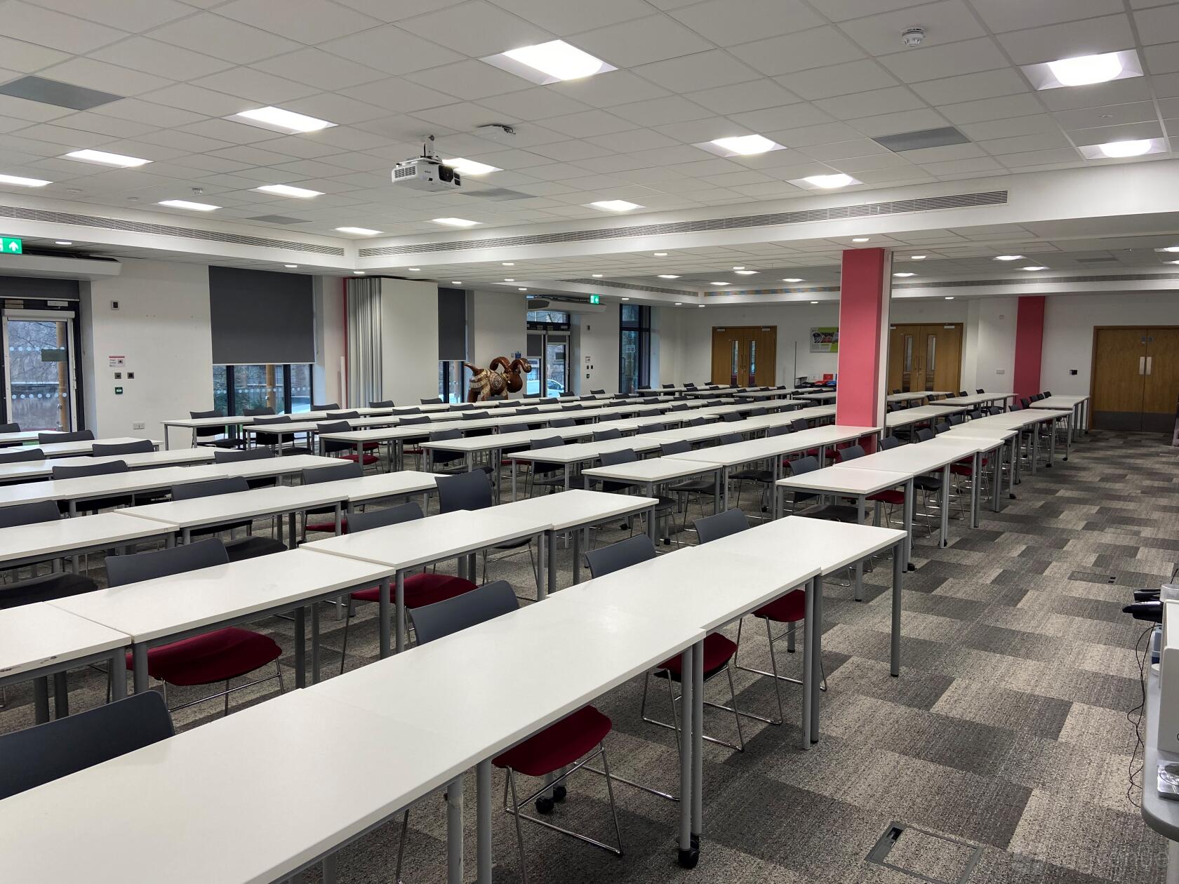 An academic event space with rows of white tables, red chairs, and a ceiling projector at Queens Building