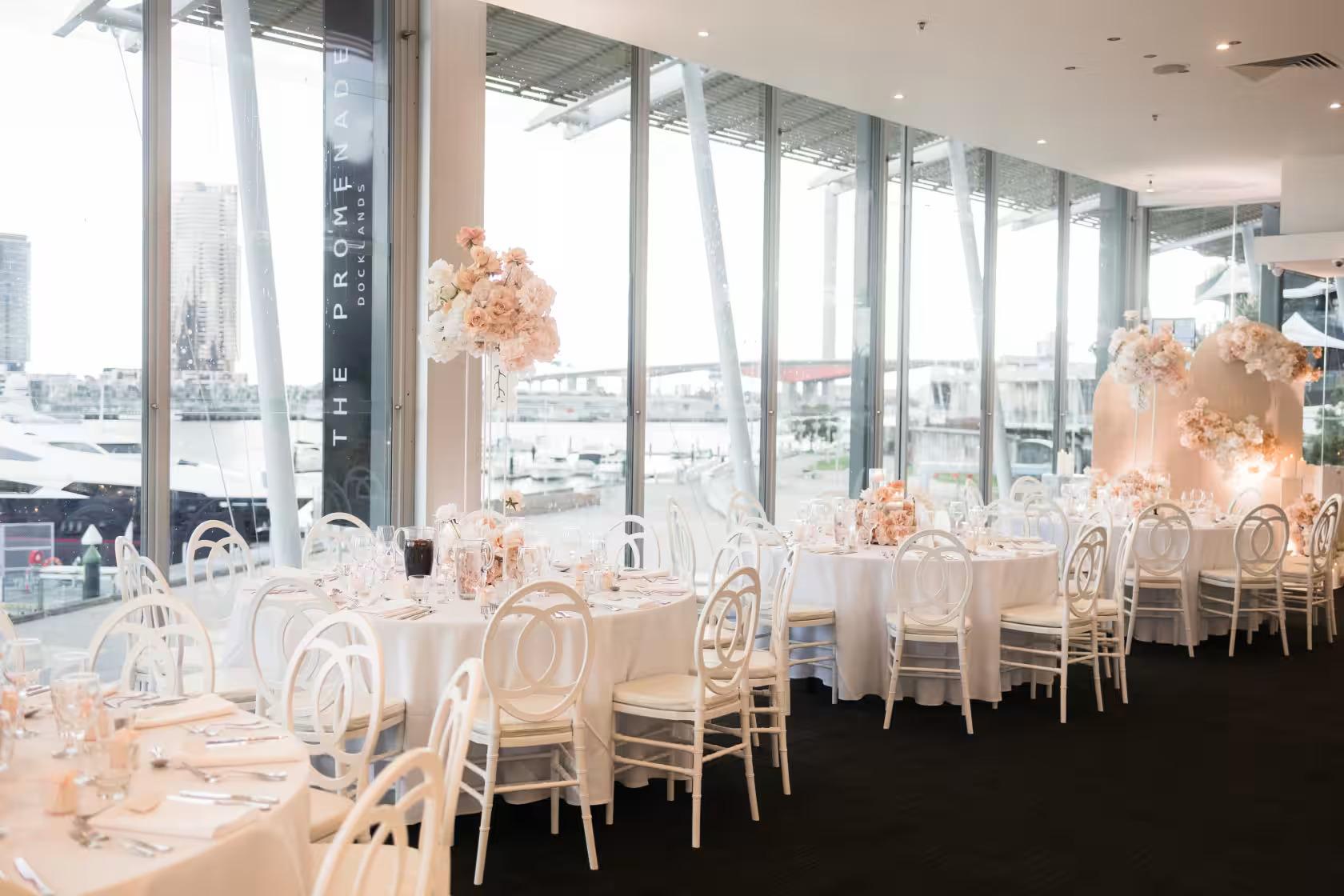 A hall with floor-to-ceiling windows, white round tables, and blush floral centerpieces at The Promenade Docklands.