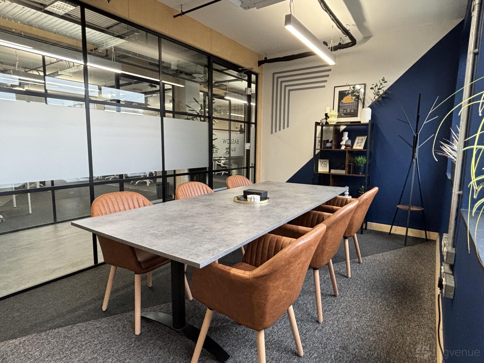 A meeting room with a concrete-style table, tan leather chairs, glass walls, and a blue accent wall at Wizu Workspace - Sheffield - Eyre Street.