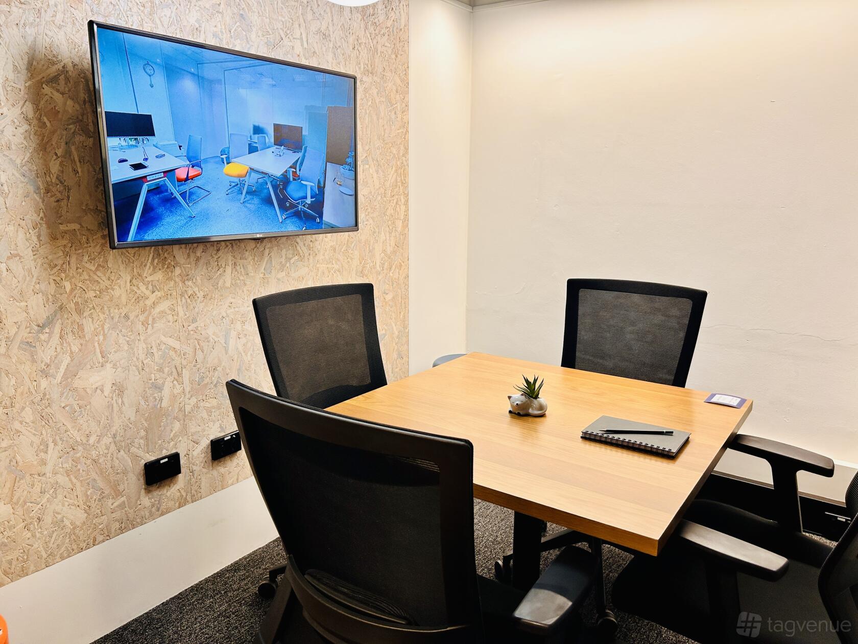 A meeting room with a wooden table, four black mesh chairs, a wall-mounted screen, and a small plant at Czar Workspace.