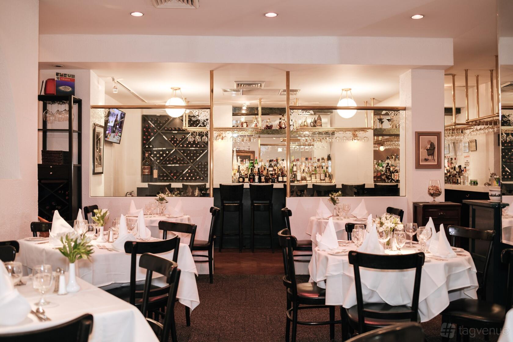 A dining room with white tablecloths, black chairs, and glass windows revealing a bar at Villa Berulia.