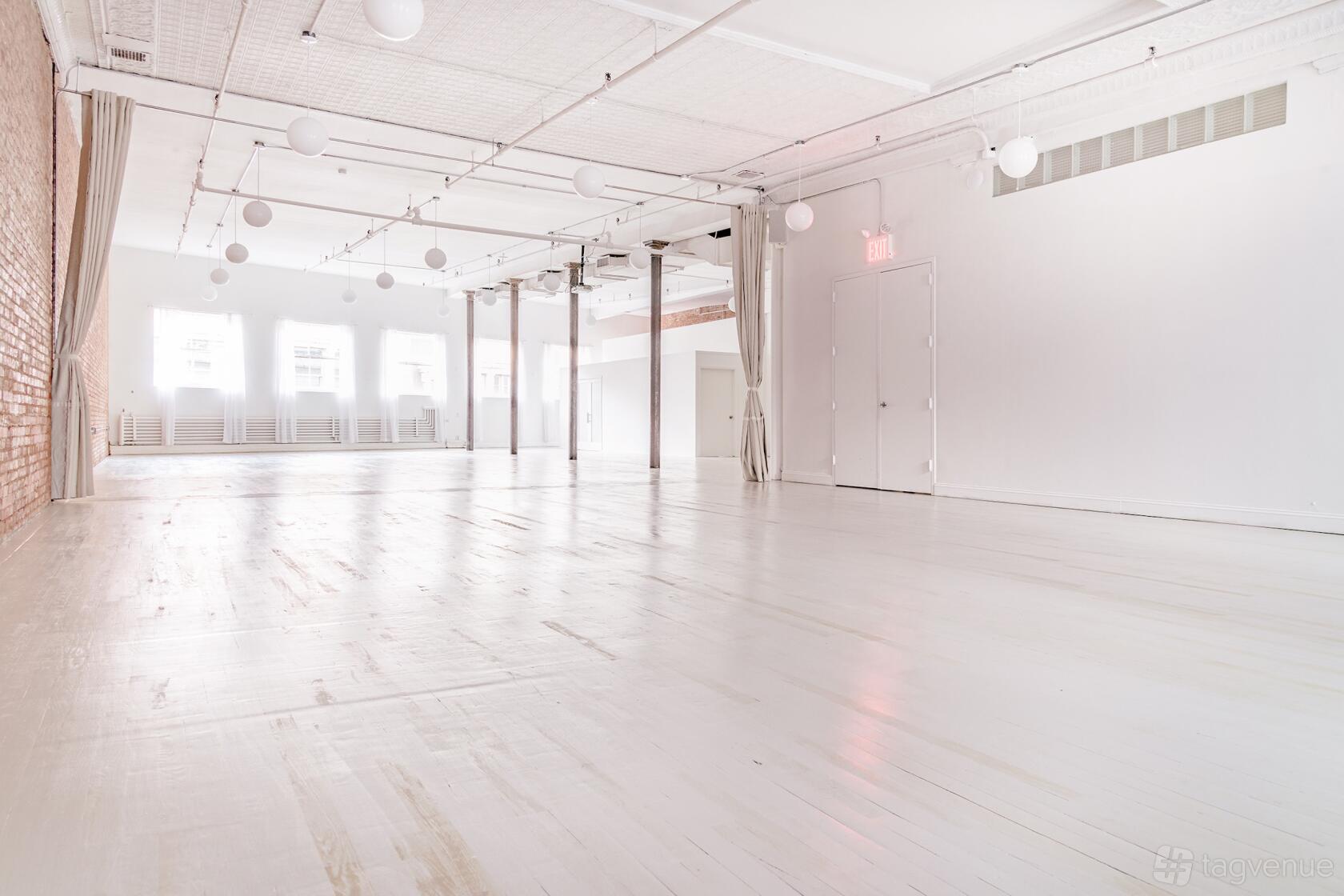 An open event space with white floors, exposed brick wall, globe pendant lights, and natural light at The Sixth Floor Loft.