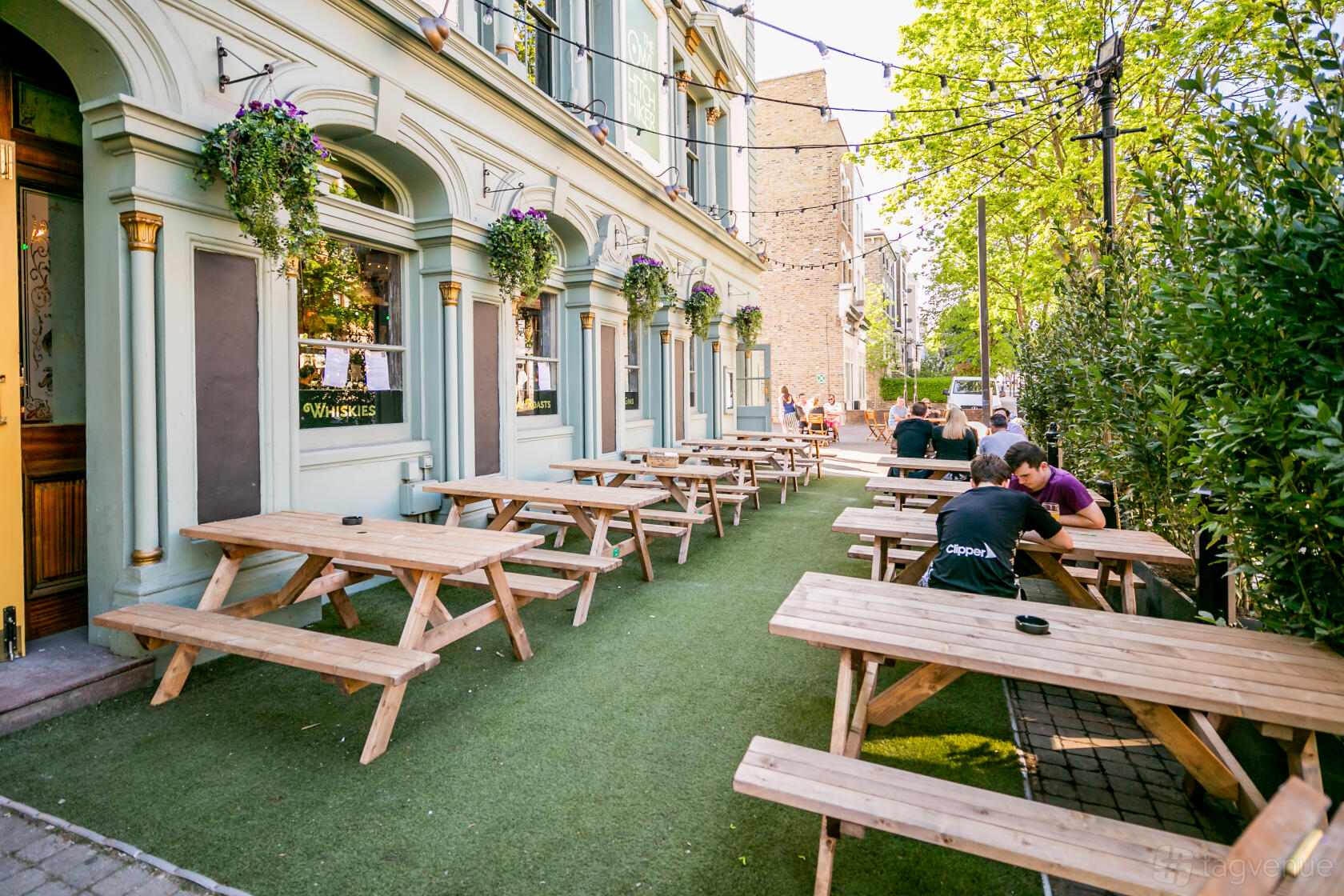 An outdoor pub area with wooden picnic tables, artificial grass, and hanging flower baskets at Owl & Hitchhiker.