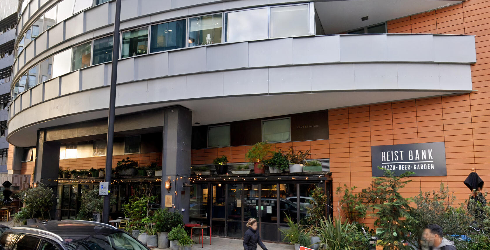 A pub with a glass frontage, orange paneling, string lights, and greenery at Heist Bank.