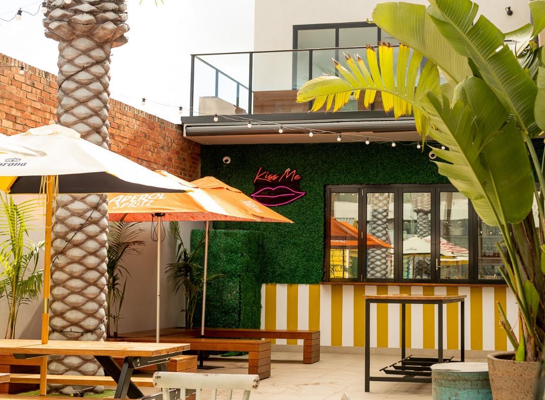 An outdoor bar space with palm trees, picnic tables, striped yellow accents, and a 'Kiss Me' neon sign at The Bungalow Beer Garden.
