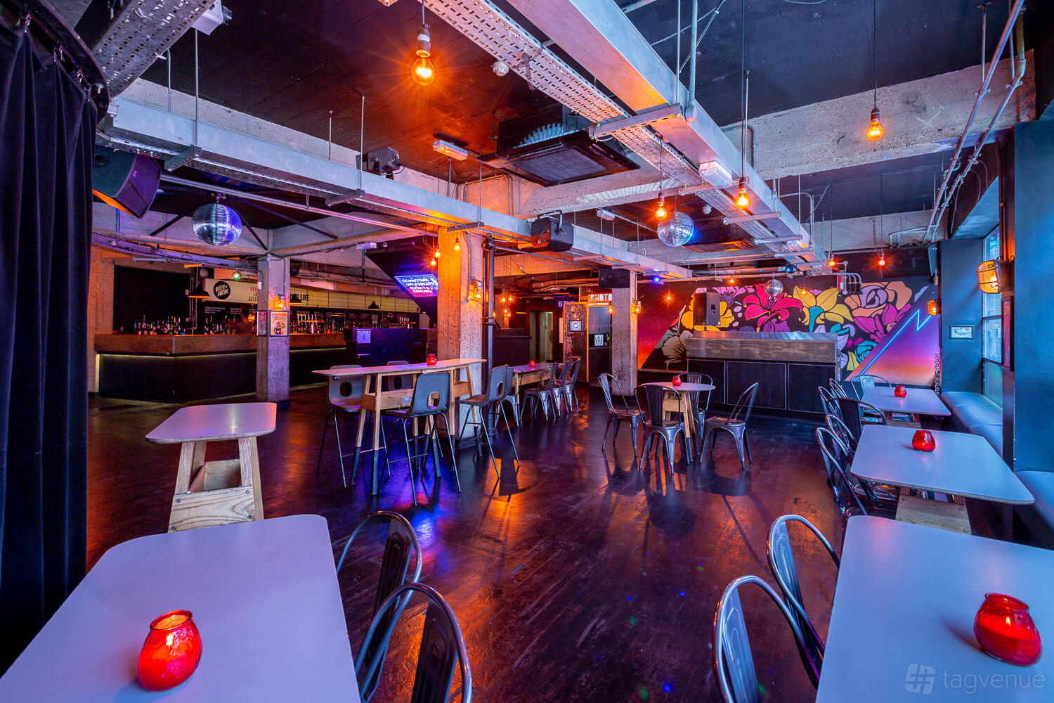 A nightclub cocktail bar with exposed ductwork, colorful graffiti wall, disco balls, and candlelit tables at Queen of Hoxton.