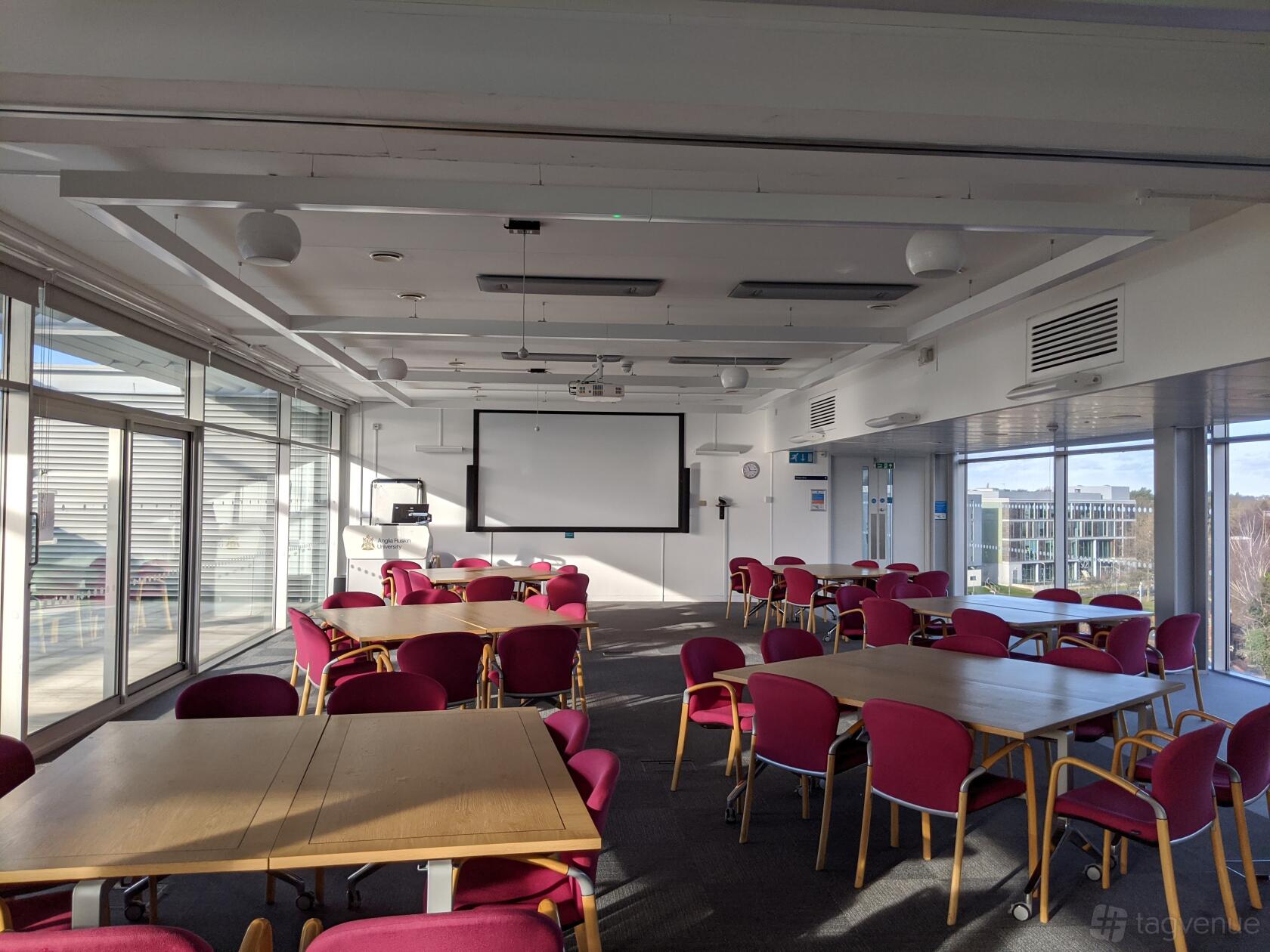 A university boardroom with glass walls, a projector screen, and cluster seating with pink chairs at ARU Venue Hire - Chelmsford.