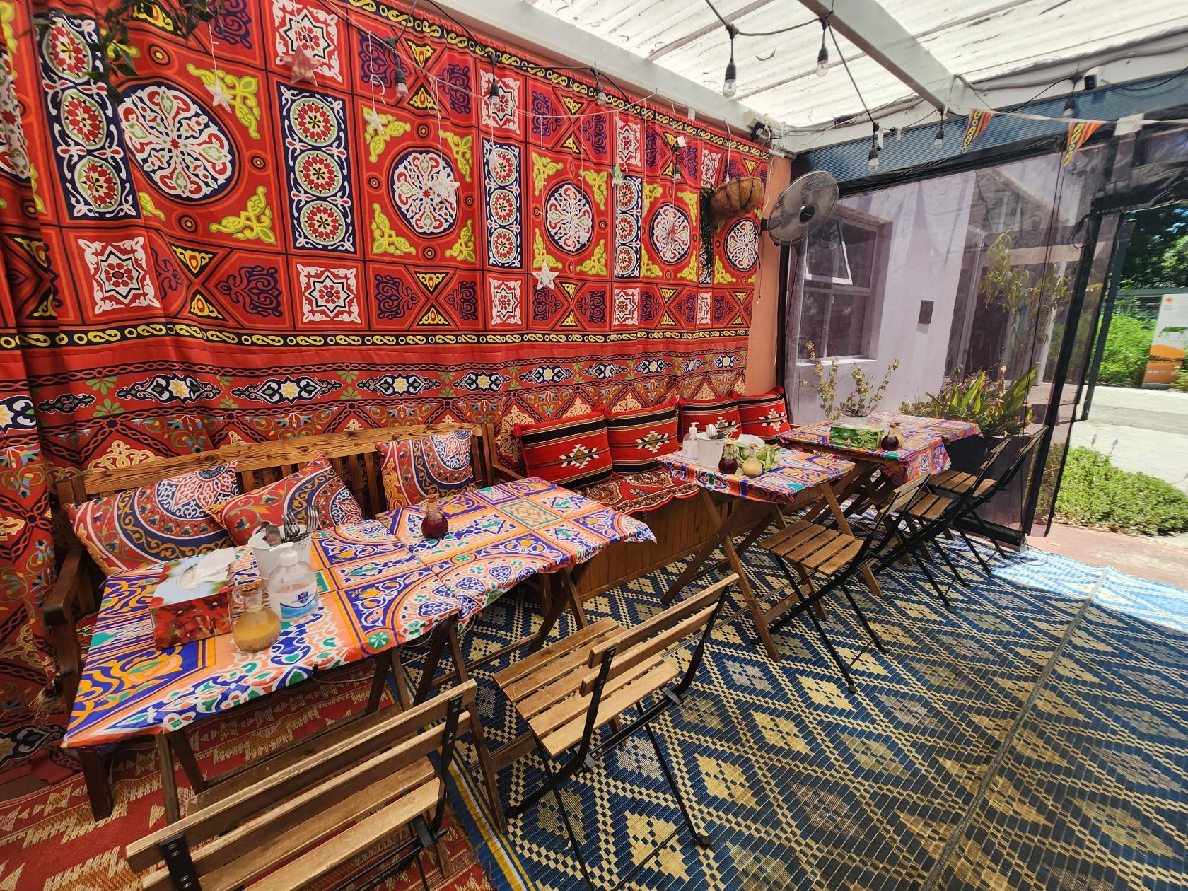A restaurant with colorful patterned tablecloths, wooden chairs, and vibrant tapestry wall at Koshari Korner.