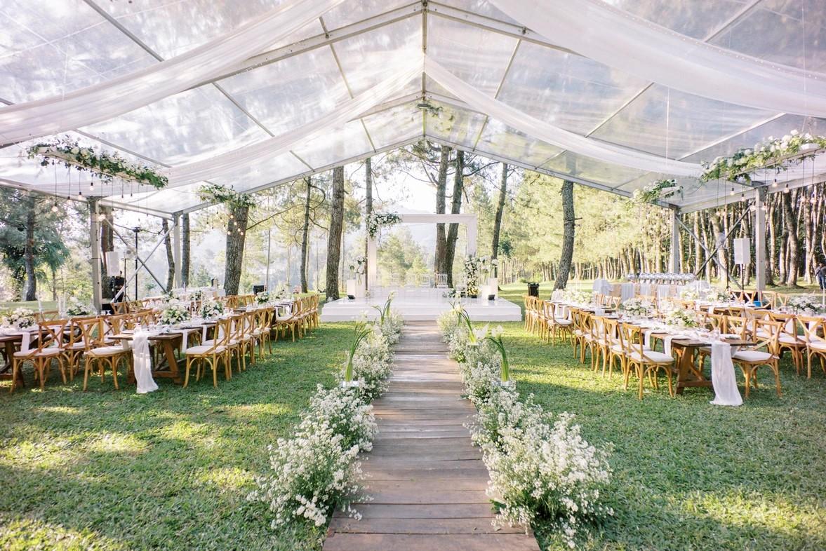 A terrace with a transparent tent, wooden walkway lined with white flowers, and banquet tables at Maceo’s Events.