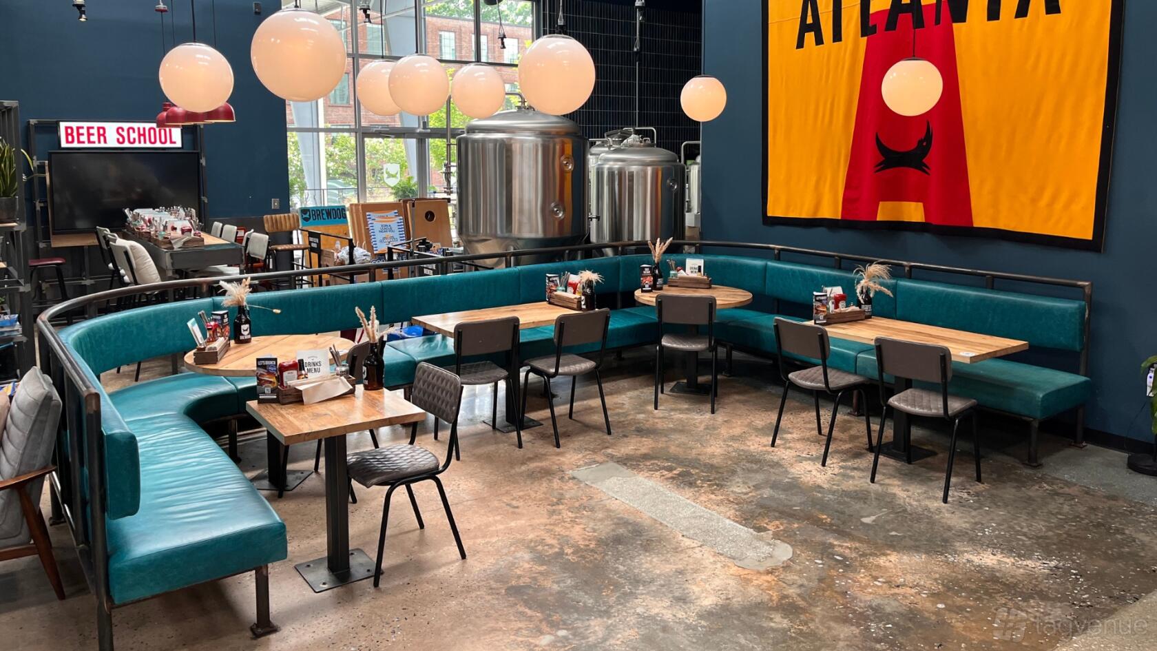 An event space in a bar with teal banquette seating, wooden tables, globe pendant lights, and brewery tanks at BrewDog Atlanta.