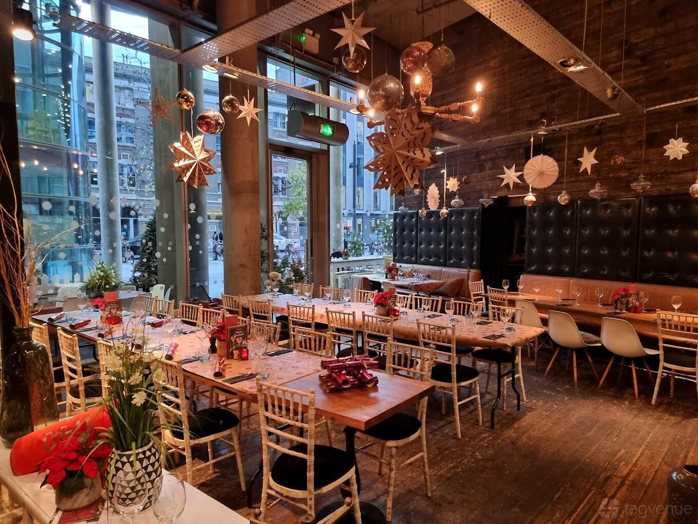 An event space in a restaurant with floor-to-ceiling windows, festive hanging decor, and banquet seating at The Refinery Bankside.