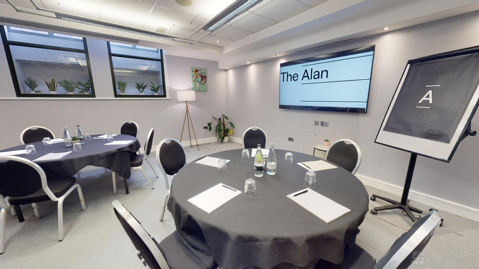 A hotel meeting room with round tables set with notepads, bottled water, and a large screen at The Alan.