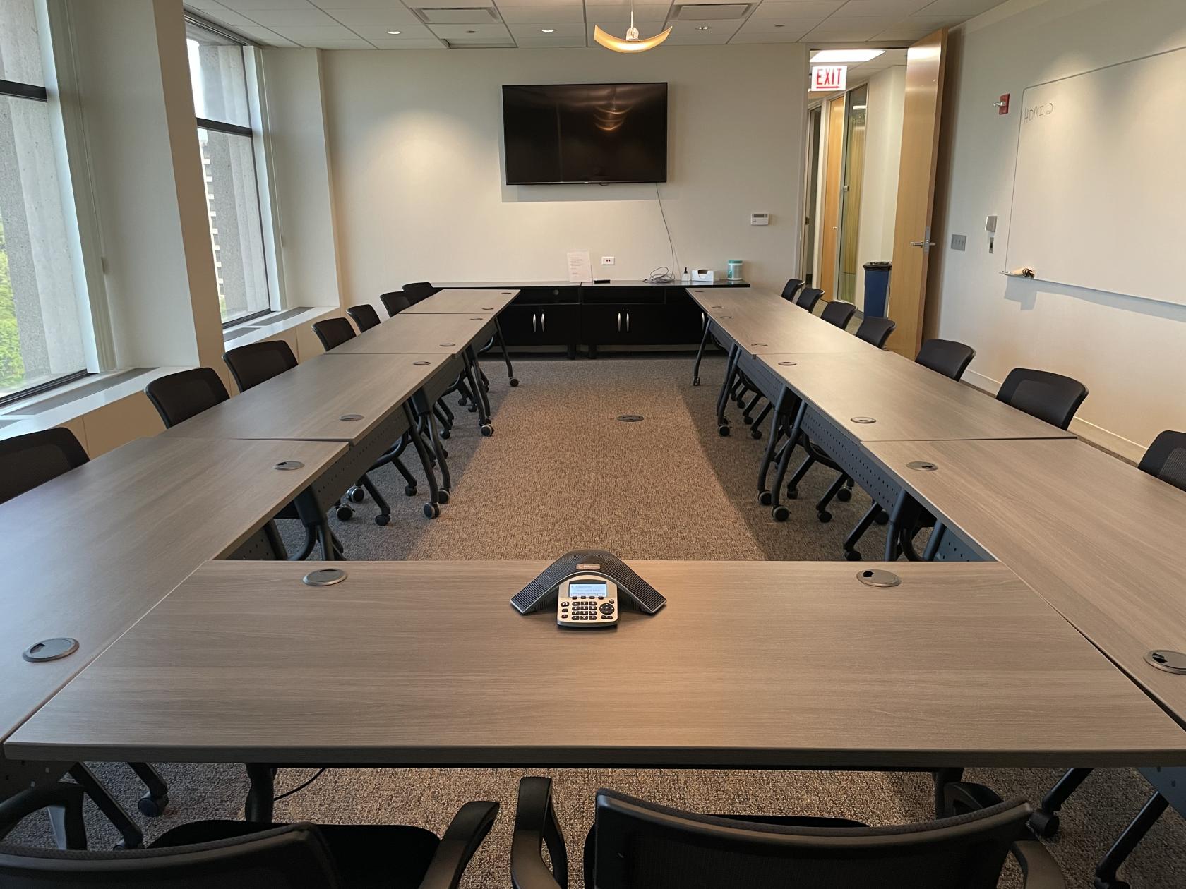 A conference room with U-shaped tables, black chairs, large windows, and a wall-mounted screen at Office Evolution O'Hare.