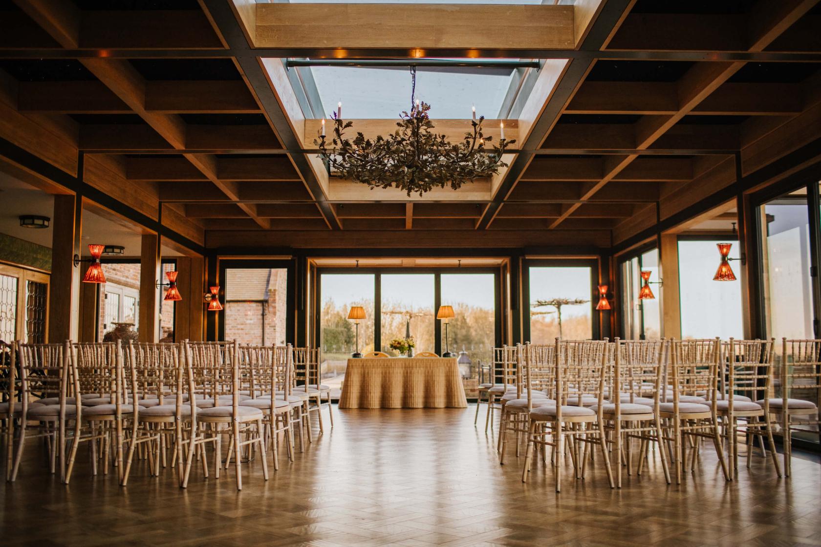 A hall with a large glass ceiling, chandelier, parquet floors, and rows of chairs facing a table at The Old Hall Ely.