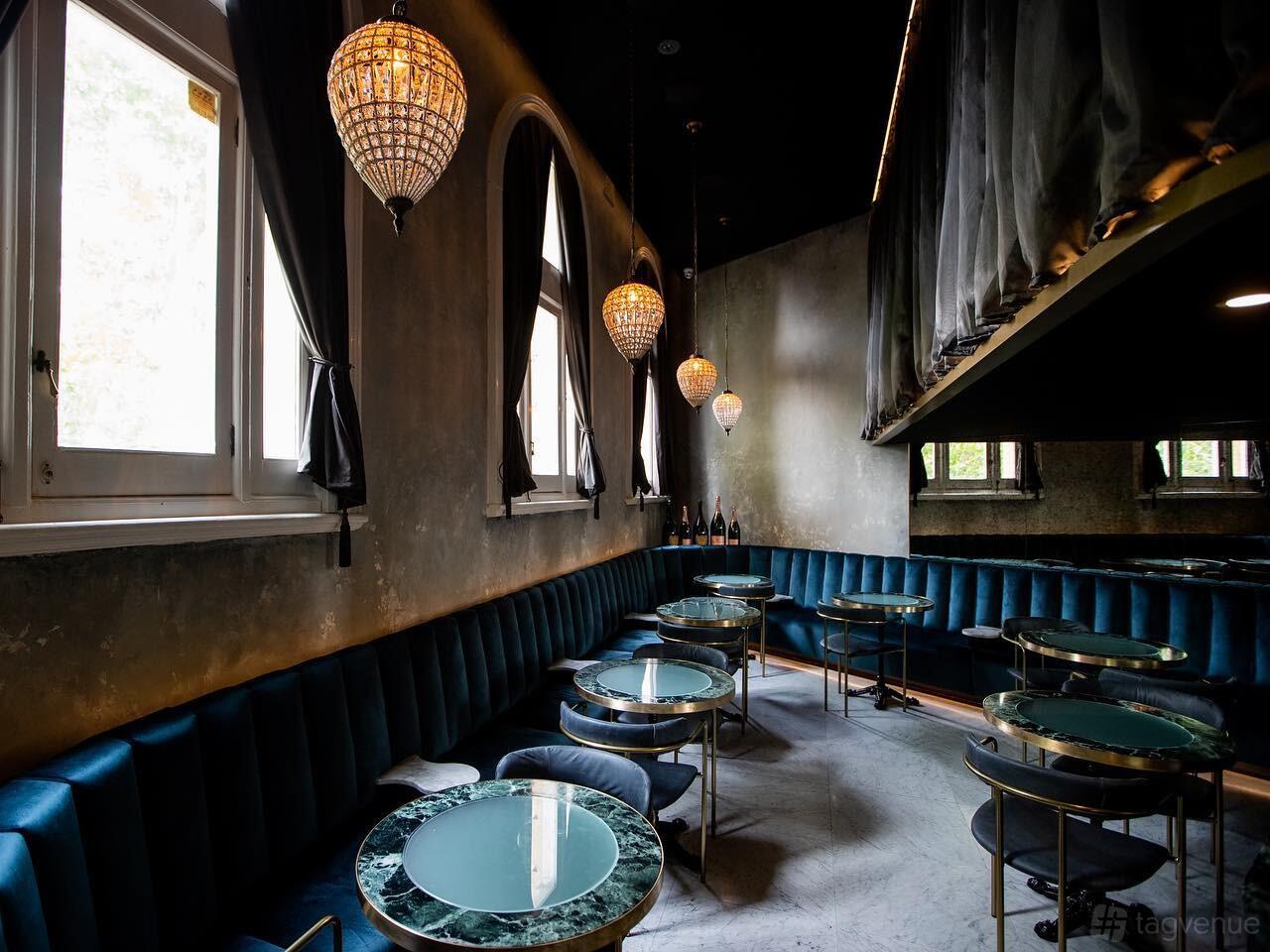 An event space in a cocktail bar with velvet banquette seating, marble-topped tables, and chandeliers at The Cat's Meow.