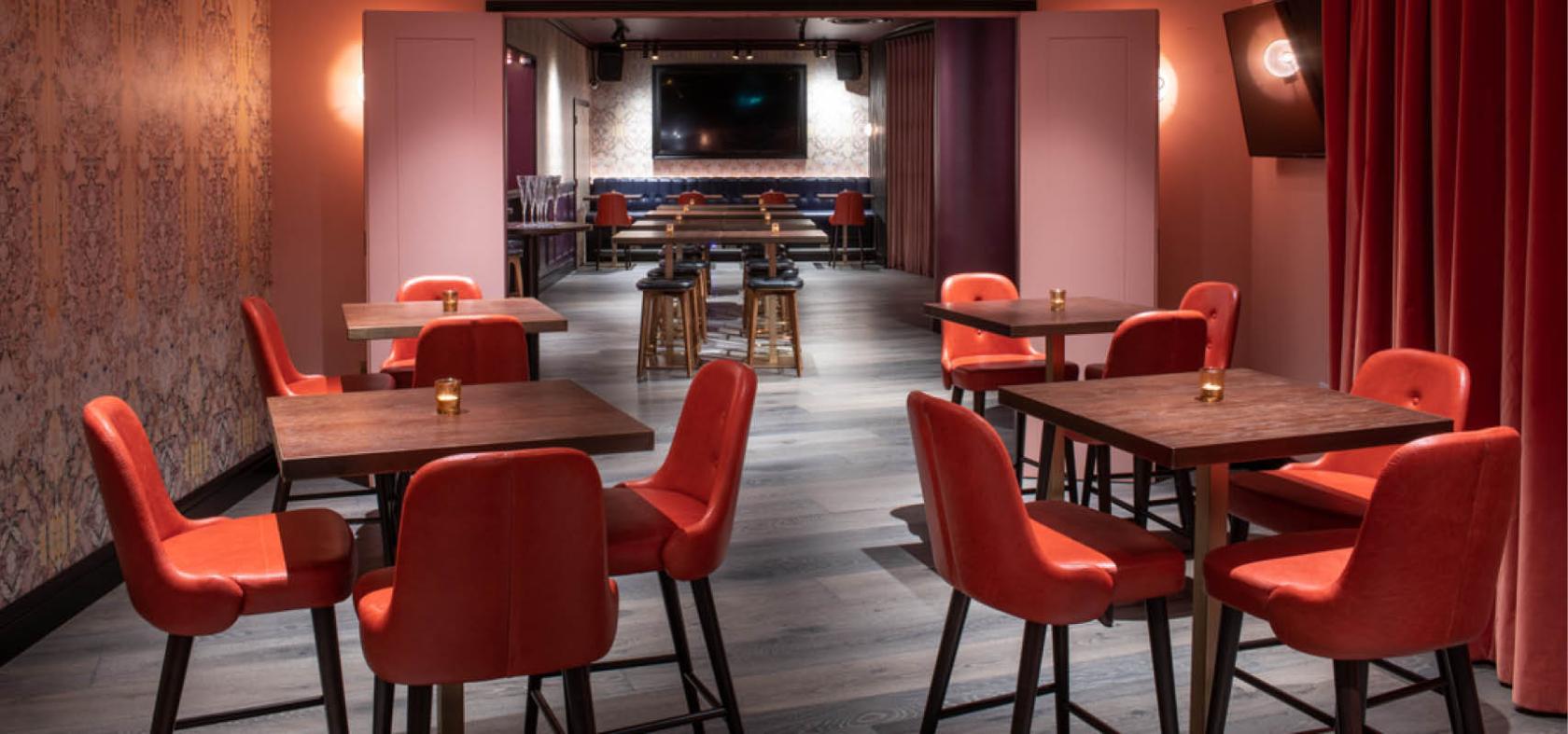 An event venue with red upholstered chairs, dark wood tables, and a large screen at Puttshack Bank.