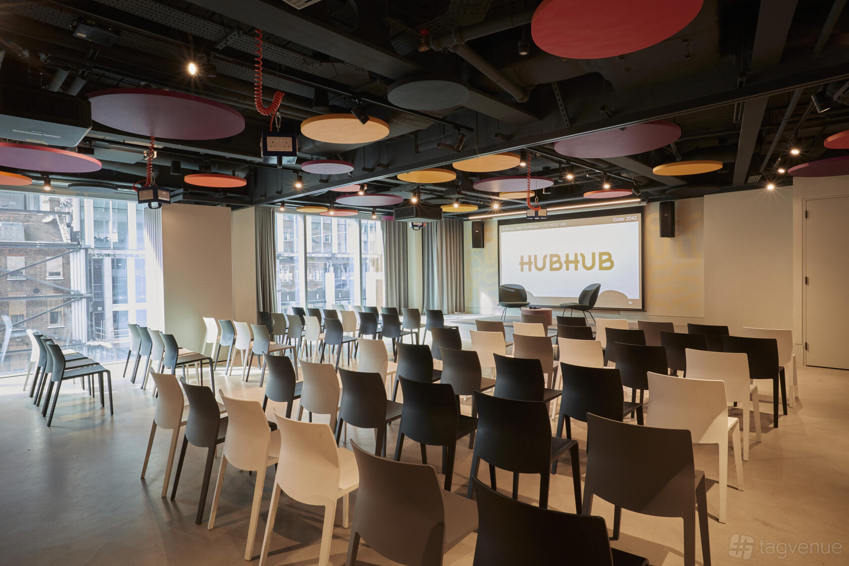An event space with rows of black and white chairs, floor-to-ceiling windows, and a large presentation screen at HubHub 20 Farringdon Street.