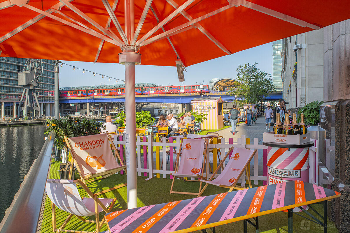 An outdoor bar venue with colorful umbrellas, canal-side seating, and lawn chairs at Fairgame Canary Wharf.