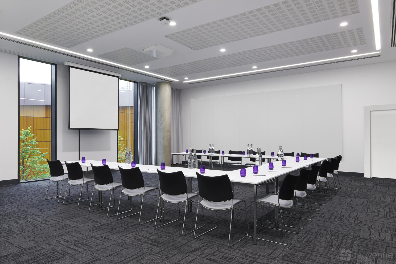 An auditorium with U-shaped tables, black chairs, water bottles, and a ceiling projector at INNSIDE Manchester.