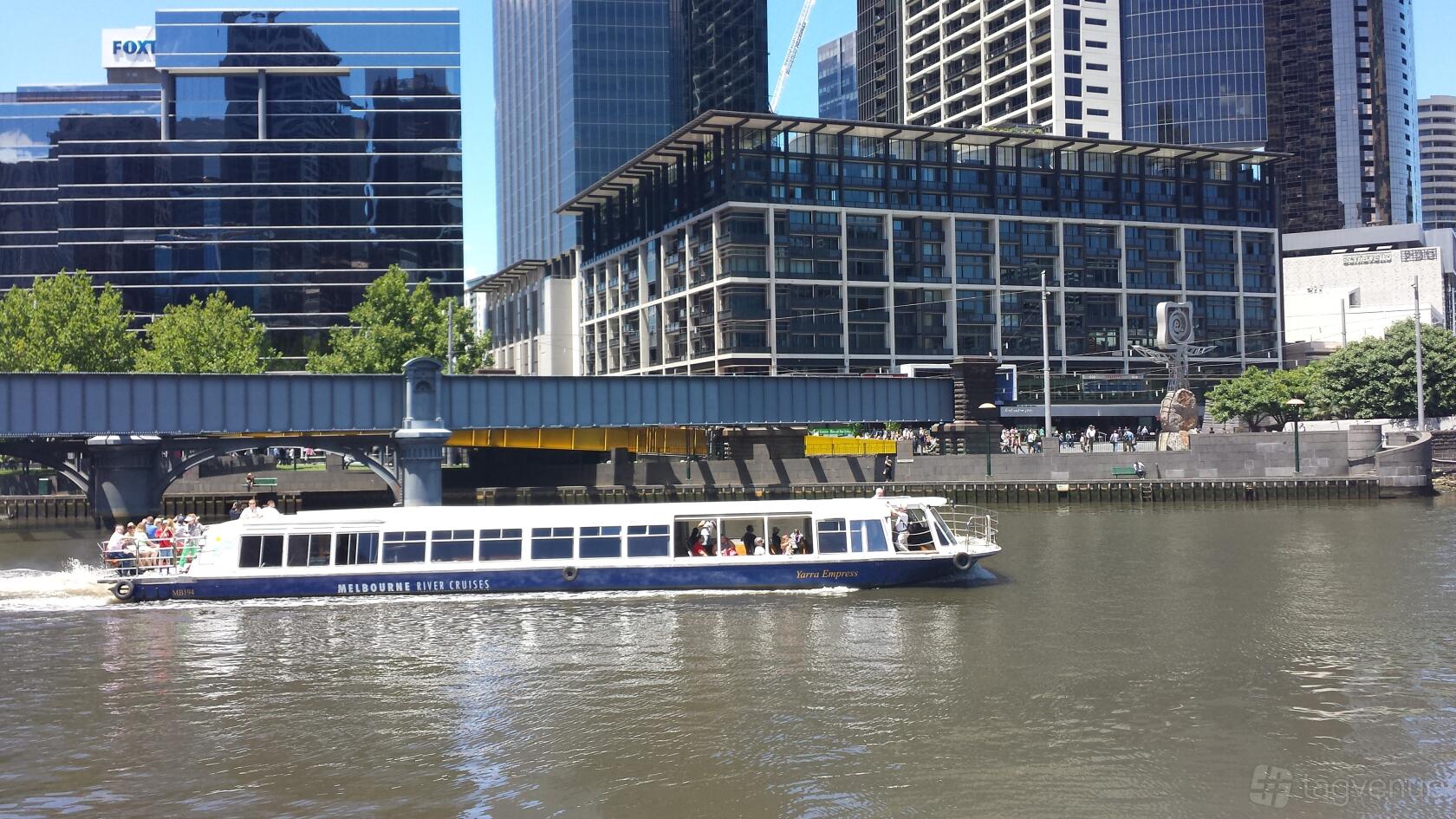 A white river cruise boat with open-air seating along the Yarra River in front of city buildings at Yarra Empress.