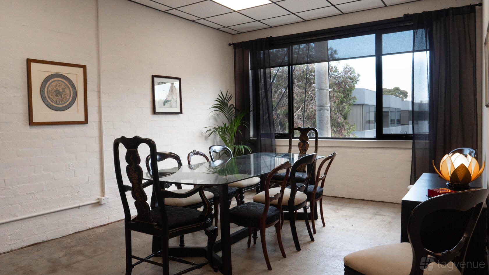 A meeting room with a glass table, eight vintage chairs, large window with sheer curtains, and framed art at Studio 14.