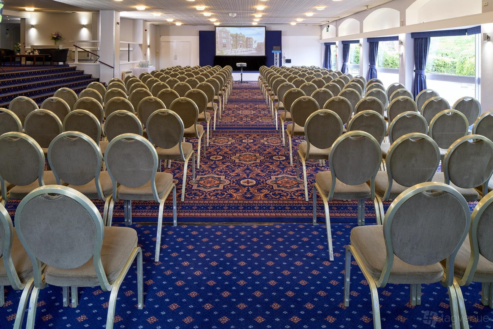 An event space with rows of round-backed chairs, patterned blue carpet, and large windows at Abbey Hotel Elgar Suite.