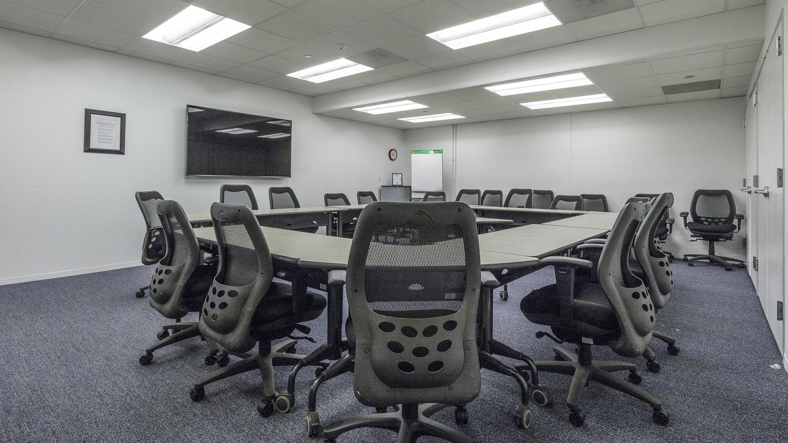 A training room with modular tables arranged in a U-shape, mesh office chairs, and a wall-mounted screen at Association Forum.