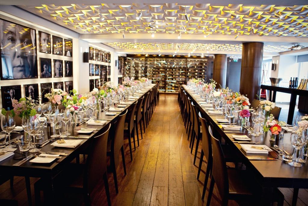 A hotel event room with long wooden tables set with glassware, floral centerpieces, and artistic wall photos at Andaz London Hotel.