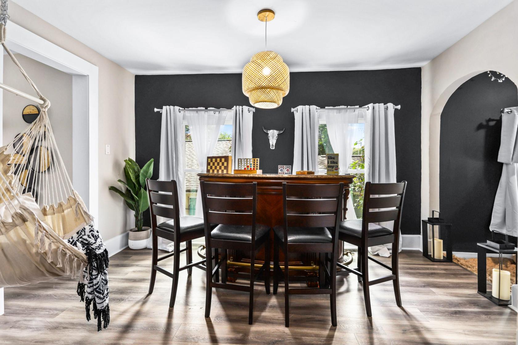 A house dining area with a round wooden table, dark accent wall, woven pendant light, and indoor hammock at 'Country Boho' 3 Bedroom Home.