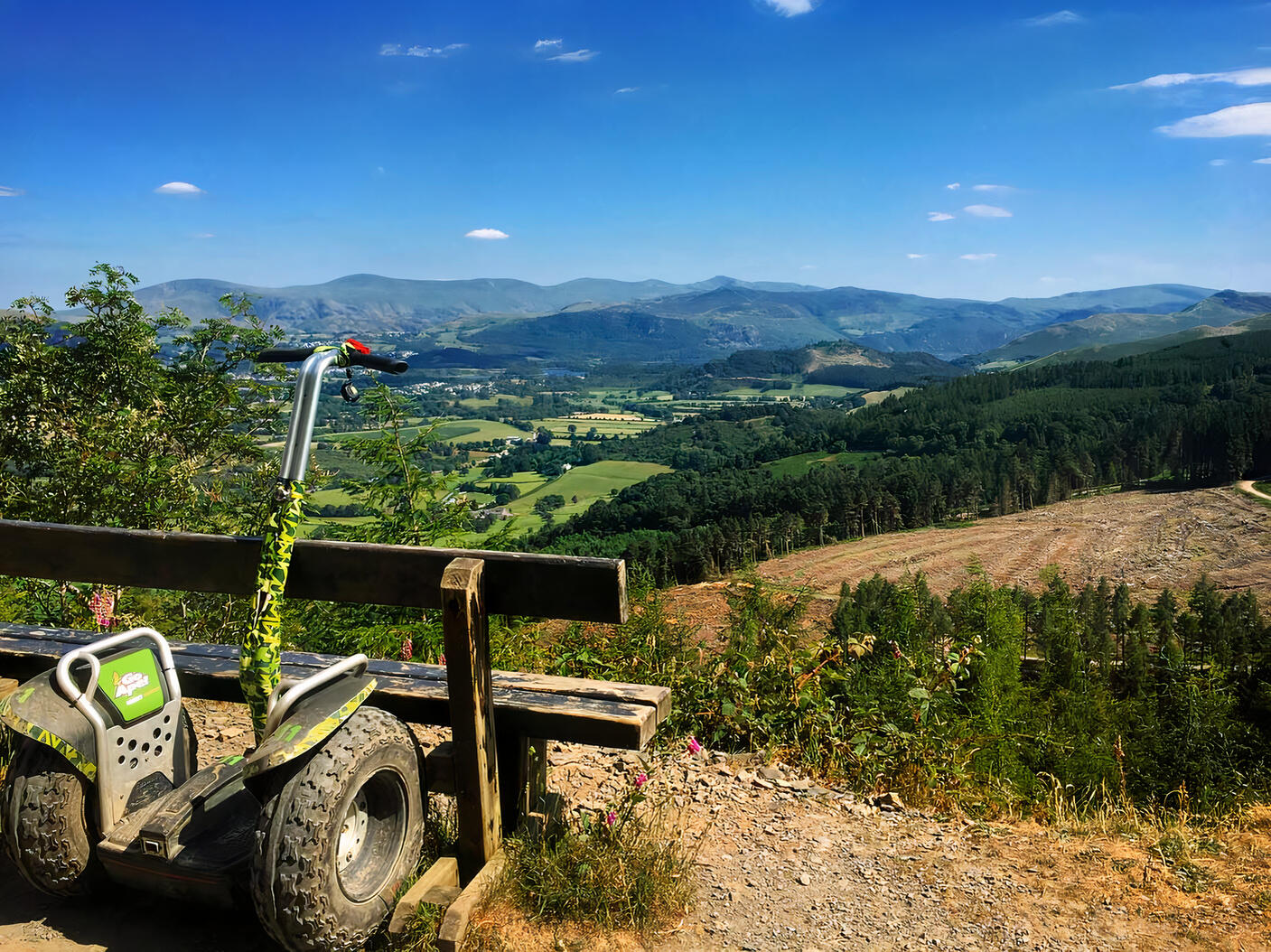 An outdoor venue with a dirt path, wooden bench, Segway, and scenic views of forests and rolling hills at Go Ape Whinlatter.