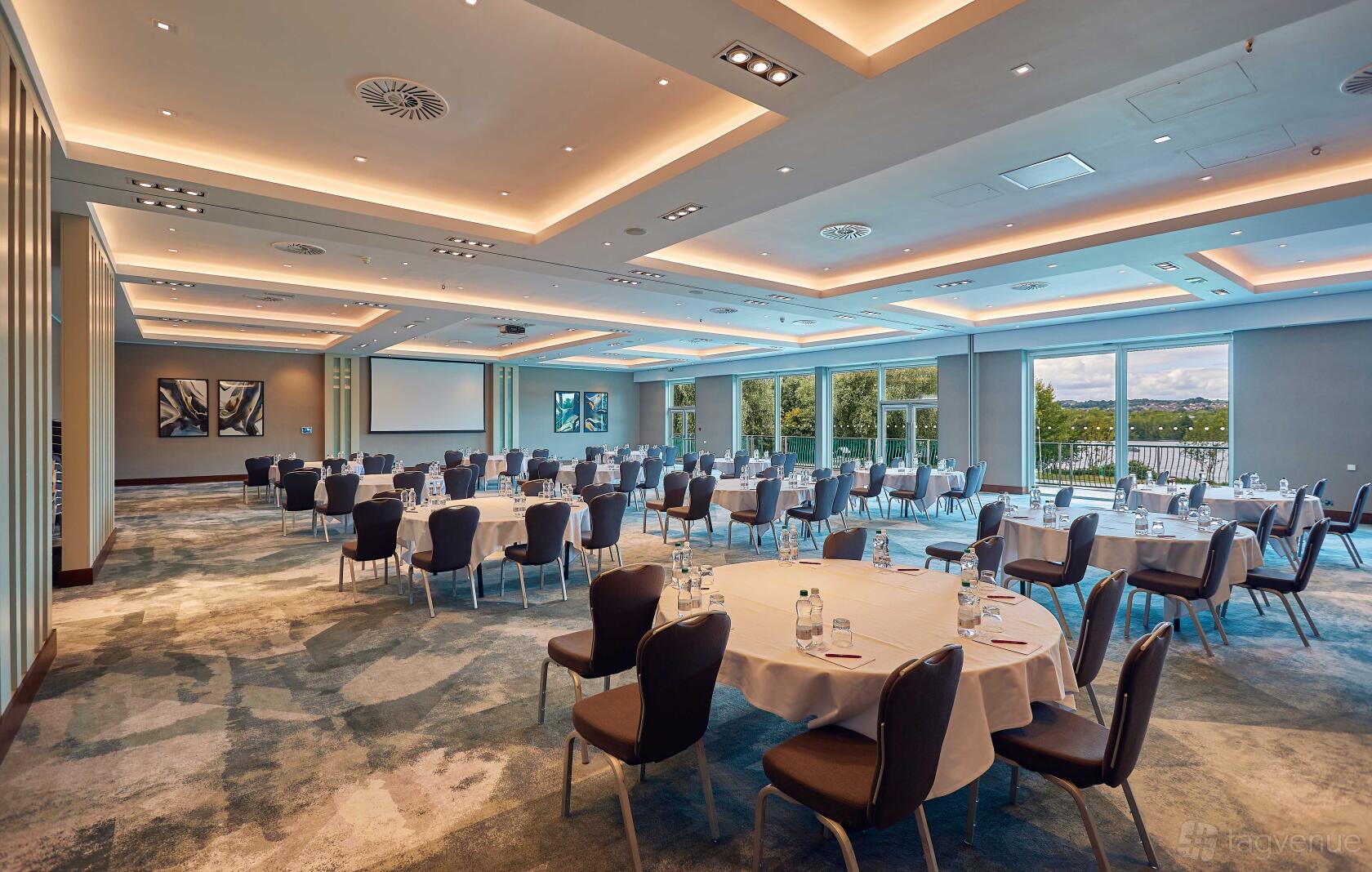 An auditorium with round tables, cushioned chairs, floor-to-ceiling windows, and projection screens at Crowne Plaza Marlow