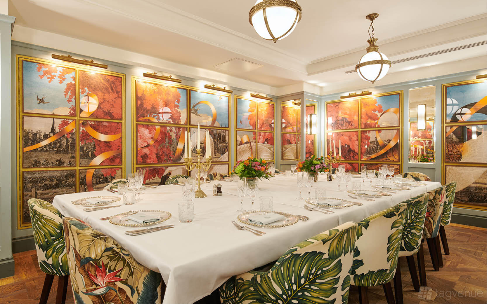 A dining room with floral-upholstered chairs, a long table set with white linens, and colorful wall murals at The Ivy Cambridge Brasserie.