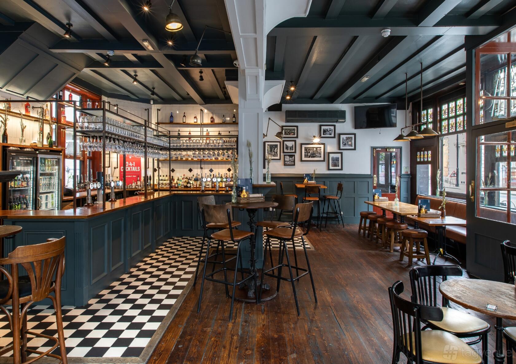 An event space in a pub with a long wooden bar, hanging glassware, checkered tile flooring, and wooden tables at Duke of Hammersmith.