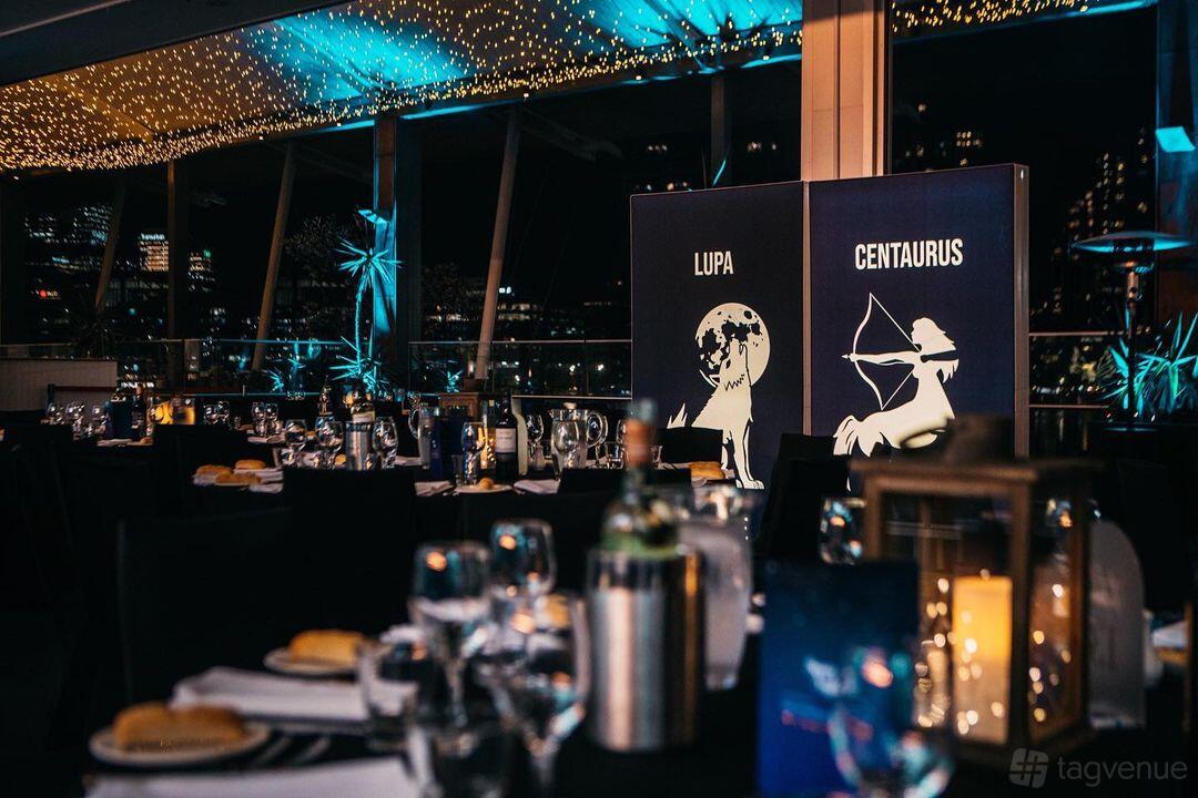 A hall with floor-to-ceiling windows, city views, string lights, and banquet tables at The Promenade Docklands.