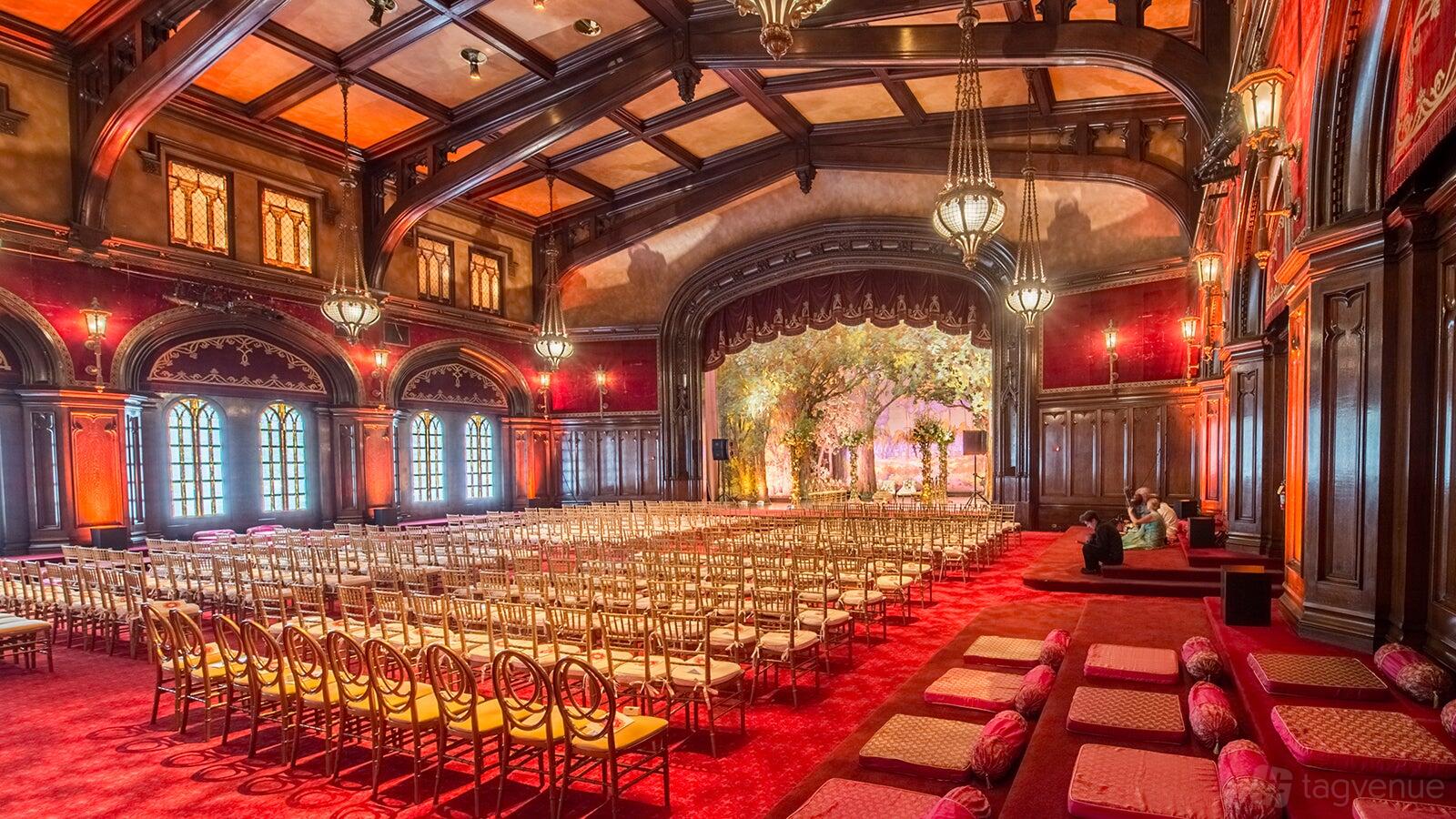 A banqueting hall with arched windows, ornate chandeliers, red carpet, and rows of gold chairs at The Lodge.