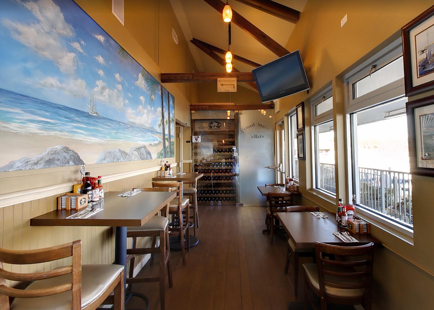 A restaurant with wooden tables, beach mural wall art, and natural light from large windows at Proud Mary's Restaurant.