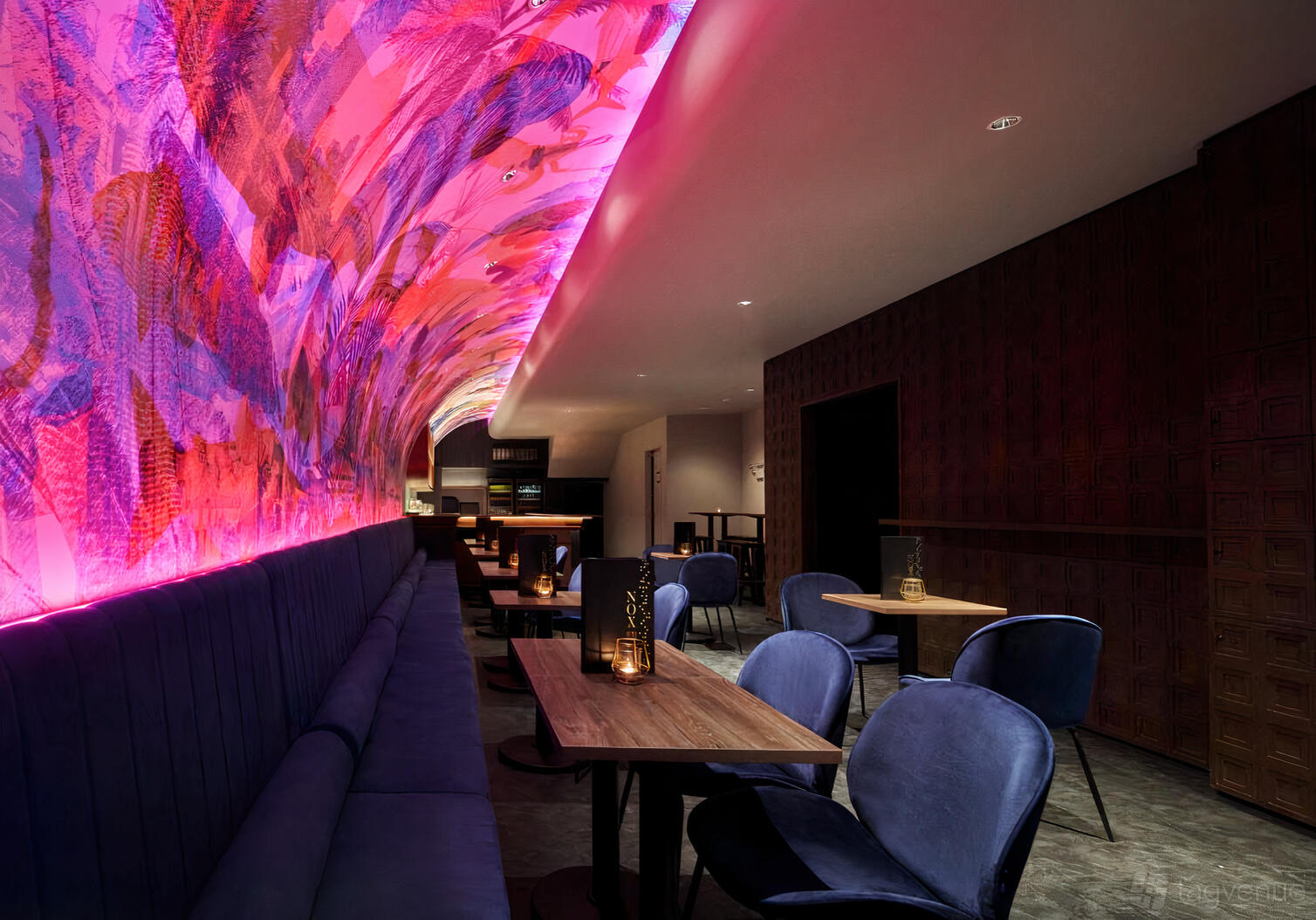 A restaurant with a curved illuminated mural ceiling, dark seating, and wooden tables at NOX - Dine in the Dark.
