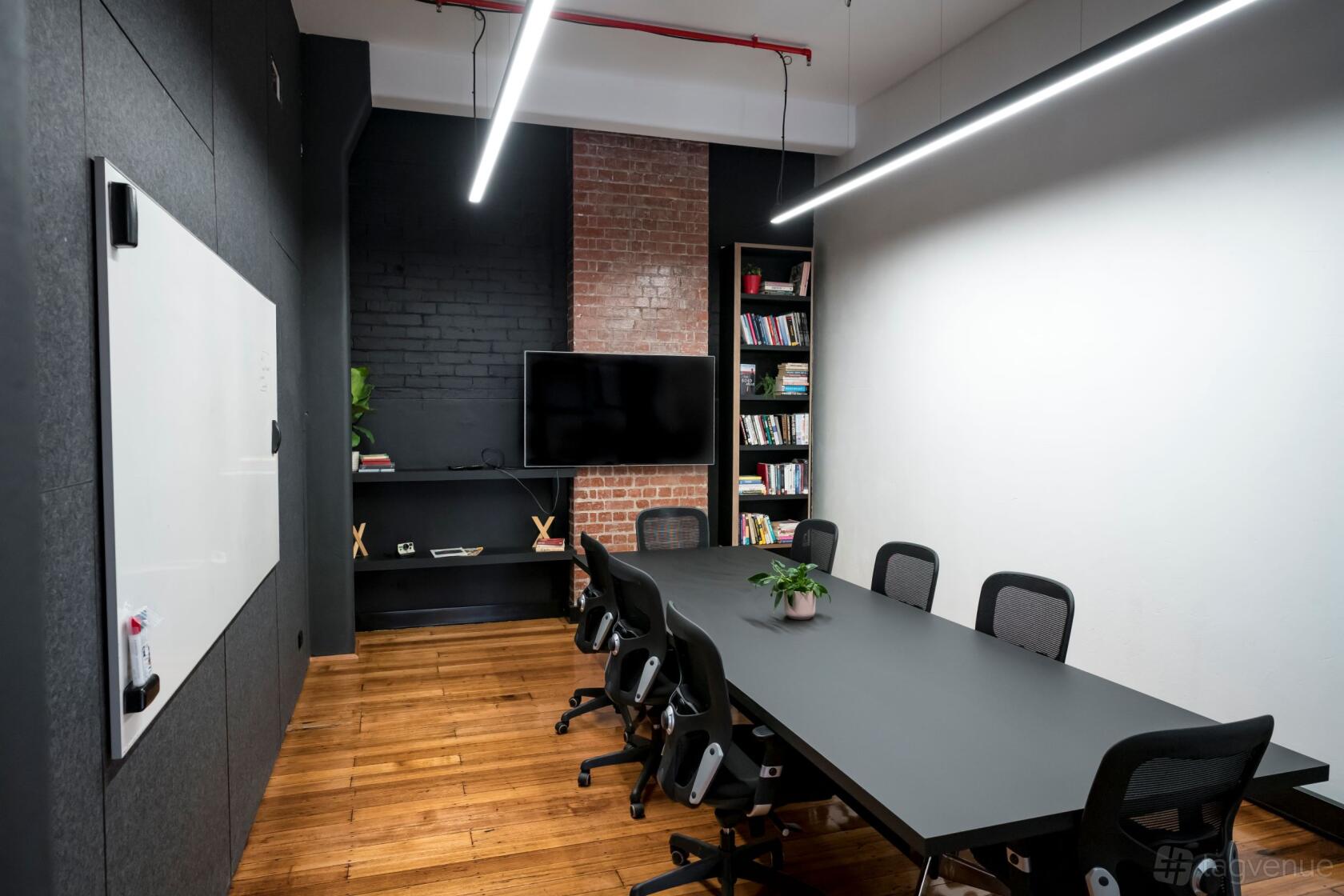 A meeting room with exposed brick wall, wall-mounted TV, bookshelves, and a long table with mesh chairs at Boiler room.