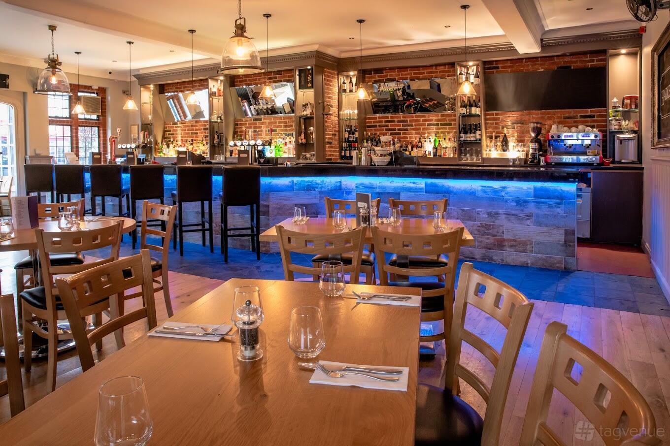 A dining room with wooden tables, matching chairs, exposed brick walls, and a backlit bar at Hadley House Restaurant & Bar.
